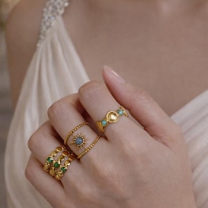 Könnte beinhalten: Eine Hand, die mehrere goldene Ringe trägt, die jeweils mit Edelsteinen verziert sind. Die Ringe weisen Designs mit grünen und türkisfarbenen Steinen sowie ein zentrales Sternmuster auf. Die Ringe sind vor einem weichen, neutralen Hintergrund platziert.