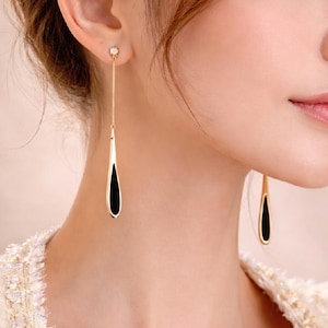 May include: Gold-coloured drop earrings with a black enamel inlay. Each earring features a small, round stud at the ear and a long, teardrop-shaped pendant. The earrings are worn by a person with light skin and brown hair.