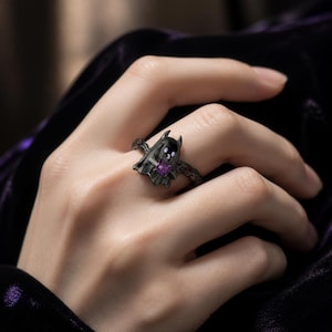 Puede incluir: Un anillo de metal negro con un diseño en forma de fantasma que presenta dos pequeños ojos blancos y una boca pequeña. La figura del fantasma está acentuada con una piedra preciosa morada de corte cuadrado en el centro y la banda es oscura.