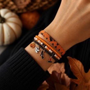 Könnte beinhalten: Ein Set aus vier gestapelten Armbändern, bestehend aus einem orangefarbenen Stoffband mit schwarzen Mustern, einer Reihe orangefarbener und weißer Perlen, einer Reihe schwarzer Perlen und einem schwarzen Perlenarmband mit Kürbis-, Geist-, Fledermaus- und Hutanhängern.