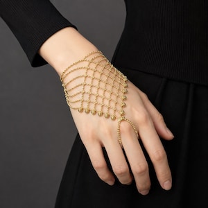 May include: A gold-toned slave bracelet featuring a delicate chain-link mesh design that covers the back of the hand. The piece includes a wrist chain and a single finger loop, with small metallic beads integrated into the grid-like structure.