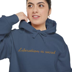 Pode incluir: Um moletom azul marinho com o texto "Liberation is sacred" em escrita dourada. O moletom tem capuz com cordão e um bolso frontal. A modelo está usando o moletom.