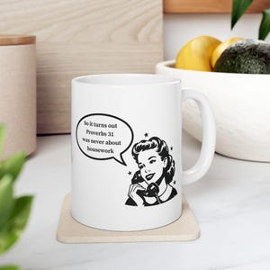 Puede incluir: Taza de cerámica blanca con un gráfico en blanco y negro de una mujer hablando por teléfono y el texto "So it turns out Proverbs 31 was never about housework." La taza tiene un asa curva y está sobre un posavasos beige.