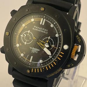 May include: A black Panerai Submersible watch with a black rubber strap. The dial features white hour markers, yellow accents, two sub-dials, and a Navy SEALs trident emblem. The bezel includes yellow numerals and the text COUNTDOWN and REG. T.M.