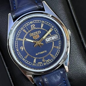 Puede incluir: Un reloj automático Seiko 5 con esfera azul marino y correa de cuero a juego. El reloj presenta agujas e indicadores dorados, una pantalla de día y fecha, y el texto "AUTOMATIC" y "JAPAN MADE". El reloj está configurado para el martes 28.