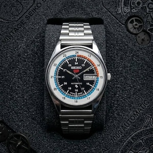 Pode incluir: Um relógio automático Seiko 5 Series vintage com pulseira de aço inoxidável. O mostrador é preto com detalhes em branco, azul e laranja. O dia e a data são exibidos às 3 horas. O relógio é apresentado em uma caixa preta.