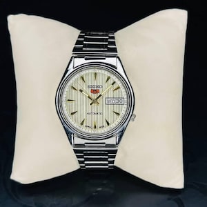 Könnte beinhalten: Eine silberne Seiko 5 Automatik-Uhr mit cremefarbenem Zifferblatt und Edelstahlarmband. Das Zifferblatt zeigt Tag und Datum an, mit goldfarbenen Zeigern und Markierungen. Die Uhr wird auf einem cremefarbenen Kissen präsentiert.