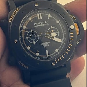 May include: A black Panerai Submersible watch with a black rubber strap, a round black dial featuring gold-toned hands, markers, and text, including the word START/STOP, FLYBACK, and REG. The dial displays a central emblem and sub-dials.