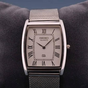 Puede incluir: Un reloj Seiko Quartz plateado con esfera rectangular y correa de malla. La esfera es blanca con números romanos y agujas negras. El reloj se muestra sobre una almohada de terciopelo gris.