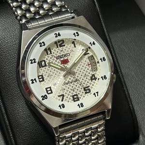 May include: Silver Seiko automatic wristwatch with a patterned white face, black numerals and hands. The watch has a date display and a stainless steel band. The watch is displayed in a black box.