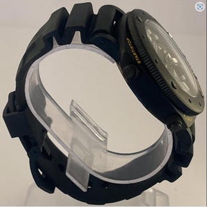 May include: A black wristwatch with a textured rubber strap and a round case. The bezel features the text START/STOP and FLY-BACK in yellow lettering. The watch has a dark dial with visible hands and markers under a clear, circular crystal face.