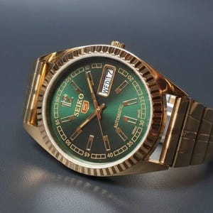 May include: A gold-coloured Seiko automatic wristwatch with a green dial. The watch has a gold band and a fluted bezel. The dial displays the day and date, with the Seiko logo clearly visible.