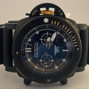 May include: A black Panerai Submersible watch with a dark blue dial, featuring a gold-colored crown, luminous hands, and a black rubber strap. The bezel includes yellow markers and the text COUNTDOWN, while the dial displays the Panerai logo.