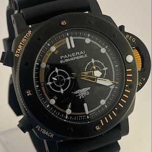 May include: A black Panerai Submersible watch with a black rubber strap. The dial features gold-toned text including START/STOP, REG., and FLYBACK, with a central trident emblem. The bezel has gold markers and the dial displays white hour indices.