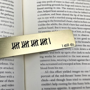 May include: A gold-colored metal bookmark with black tally marks and the words "I still do." The bookmark is rectangular and lies on top of an open book with printed text.
