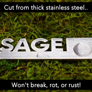 May include: A silver metal sign with the word "SAGE" cut out. The sign is on a bed of green moss. The text "Cut from thick stainless steel... Won't break, rot, or rust!" is visible above and below the sign.