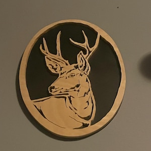 May include: A wooden deer head silhouette in a circular frame. The deer is light brown, set against a dark background, and the frame is a lighter shade of brown. The artwork is mounted on a gray wall.