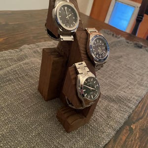 May include: A wooden watch stand displaying three wristwatches with silver bands and dark watch faces. The stand is made of dark brown wood and sits on a gray fabric surface.