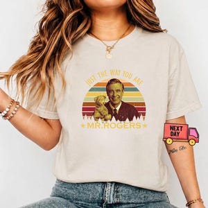May include: Beige t-shirt featuring a retro design with the text "JUST THE WAY YOU ARE" above an image of Mr. Rogers and a teddy bear. The design includes a colorful sunset and stars. The shirt is a crew neck.