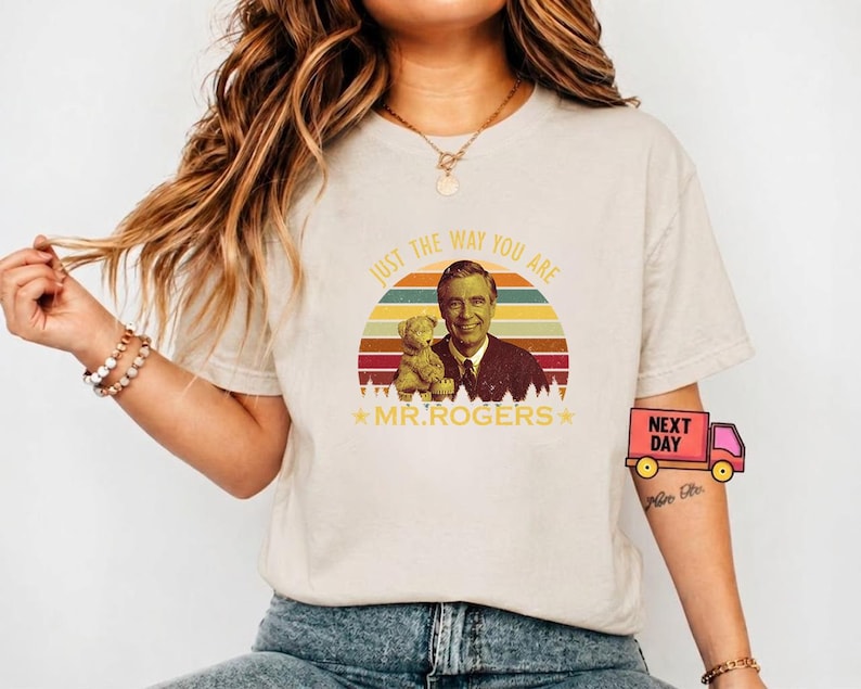 May include: Beige t-shirt featuring a retro design with the text "JUST THE WAY YOU ARE" above an image of Mr. Rogers and a teddy bear. The design includes a colorful sunset and stars. The shirt is a crew neck.