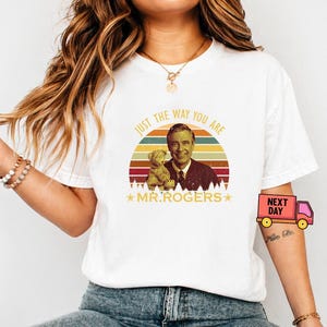 May include: White t-shirt featuring a graphic of Mr. Rogers with a teddy bear, set against a sunset design. The text "JUST THE WAY YOU ARE" and "MR. ROGERS" are also displayed. The shirt is a casual wear item.