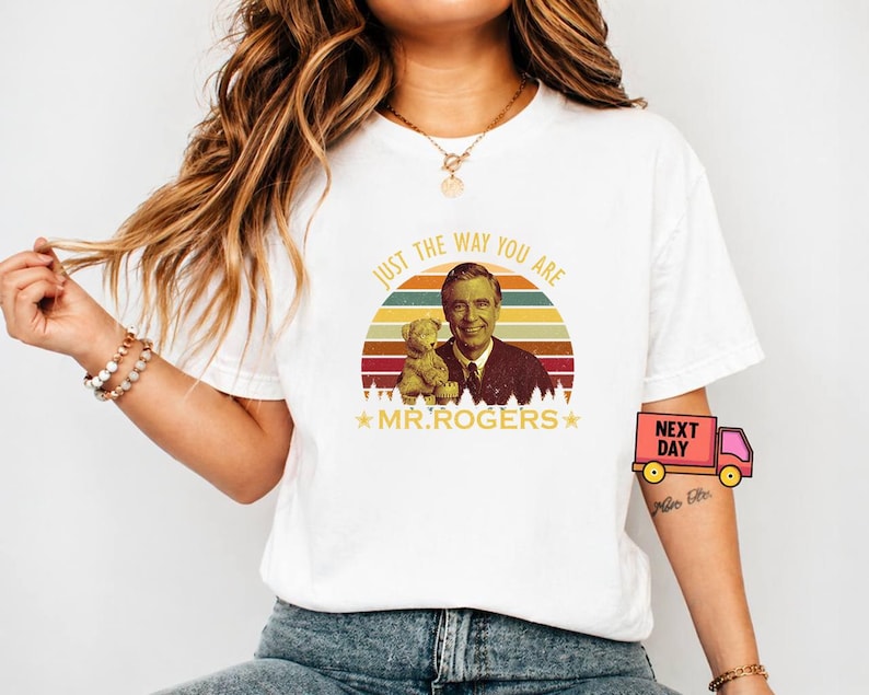 May include: White t-shirt featuring a graphic of Mr. Rogers with a teddy bear, set against a sunset design. The text "JUST THE WAY YOU ARE" and "MR. ROGERS" are also displayed. The shirt is a casual wear item.