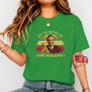 May include: Green t-shirt featuring a retro design with Fred Rogers and a teddy bear, set against a sunset backdrop. The text reads "JUST THE WAY YOU ARE" and "MR. ROGERS".