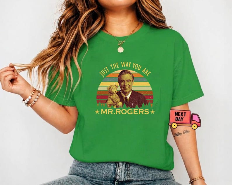 May include: Green t-shirt featuring a retro design with Fred Rogers and a teddy bear, set against a sunset backdrop. The text reads "JUST THE WAY YOU ARE" and "MR. ROGERS".