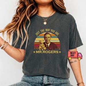 May include: Dark gray t-shirt featuring a vintage-style graphic of Mr. Rogers holding a teddy bear, set against a sunset design. The text "JUST THE WAY YOU ARE" and "MR. ROGERS" are included. The shirt is a casual wear item.