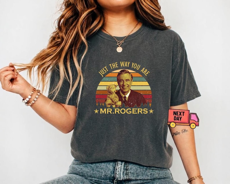May include: Dark gray t-shirt featuring a vintage-style graphic of Mr. Rogers holding a teddy bear, set against a sunset design. The text "JUST THE WAY YOU ARE" and "MR. ROGERS" are included. The shirt is a casual wear item.