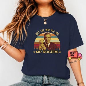 May include: Navy blue t-shirt featuring a retro design of Mr. Rogers with a teddy bear, set against a sunset backdrop. The text "JUST THE WAY YOU ARE" and "MR. ROGERS" are included in the design. A vintage-style tee.
