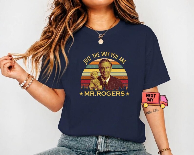 May include: Navy blue t-shirt featuring a retro design of Mr. Rogers with a teddy bear, set against a sunset backdrop. The text "JUST THE WAY YOU ARE" and "MR. ROGERS" are included in the design. A vintage-style tee.