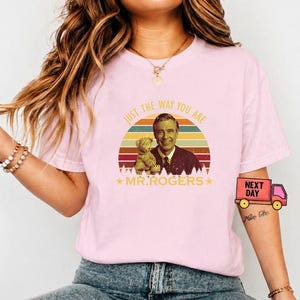 May include: A light pink t-shirt featuring a graphic of Mr. Rogers with a teddy bear, set against a retro sunset design. The text "JUST THE WAY YOU ARE" and "MR. ROGERS" are also printed on the shirt. The shirt is a casual wear item.
