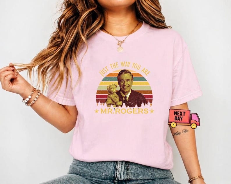 May include: A light pink t-shirt featuring a graphic of Mr. Rogers with a teddy bear, set against a retro sunset design. The text "JUST THE WAY YOU ARE" and "MR. ROGERS" are also printed on the shirt. The shirt is a casual wear item.