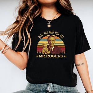 May include: Black t-shirt featuring a retro design with the text "JUST THE WAY YOU ARE" above an image of Mr. Rogers and a teddy bear, set against a sunset backdrop. The words "MR. ROGERS" are below the image.