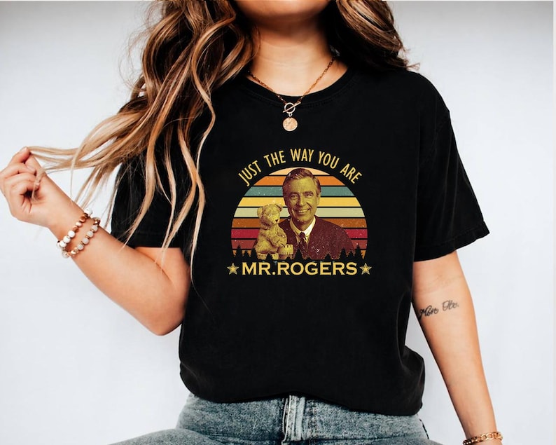 May include: Black t-shirt featuring a retro design with the text "JUST THE WAY YOU ARE" above an image of Mr. Rogers and a teddy bear, set against a sunset backdrop. The words "MR. ROGERS" are below the image.