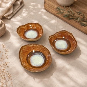 May include: Three small, round ceramic bowls with irregular, organic edges. Each bowl features a deep, glossy amber-brown glaze on the exterior and rim, transitioning into a smooth, speckled light-blue and white center basin.