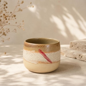 May include: A ceramic cup with a rounded shape and a speckled, textured surface. The exterior features horizontal bands of tan, cream, and light brown glaze, accented by a single, diagonal, hand-painted brushstroke in a deep reddish-pink color.