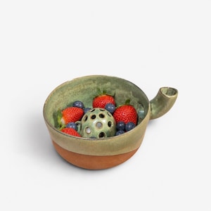May include: A round ceramic bowl with a side handle, featuring a green glazed interior and a natural terracotta exterior. Inside the bowl sits a small, perforated green ceramic dome surrounded by fresh red strawberries and dark blue blueberries.