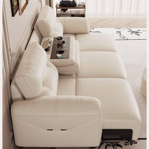 Puede incluir: Sofá seccional color crema con una mesa auxiliar incorporada. El sofá presenta un diseño moderno con una textura suave similar al cuero. La mesa auxiliar tiene una superficie oscura con portavasos y una estación de carga. El sofá está sobre una alfombra estampada.