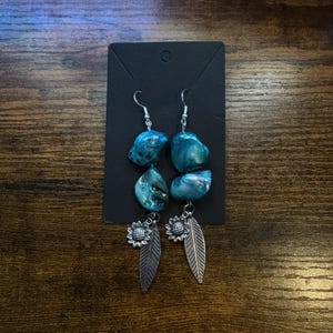 May include: A pair of dangle earrings featuring iridescent blue-green stones. Each earring has two stones, a silver sunflower, and a silver feather charm. The earrings are displayed on a black earring card against a wooden background.