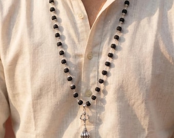 May include: A black beaded necklace with a silver pendant. The necklace features dark, round beads strung together with silver links. The pendant has a detailed, ornate design with a silver finish. The necklace is worn over a light-colored button-down shirt.