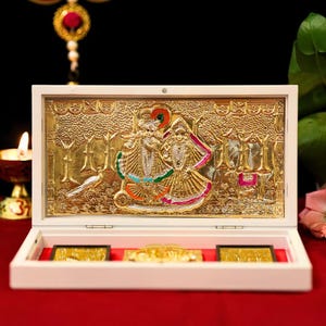 May include: A gold-colored, ornate relief sculpture of two figures in a forest scene, presented in a white box with red lining. The figures are adorned with colorful details. The box contains additional gold-colored decorative elements. A peacock is also visible in the scene.