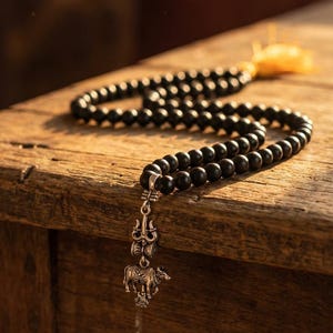 May include: Two strands of black beaded prayer beads with a silver pendant featuring a trident and an elephant. The beads rest on a weathered wooden surface, with a yellow tassel visible in the background.