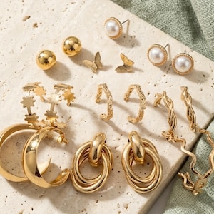 May include: A collection of gold-toned earrings displayed on a textured surface. The assortment includes stud earrings with pearls, ball studs, butterfly-shaped earrings, hoop earrings, and oval drop earrings. The earrings vary in design and size.