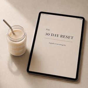 May include: A digital tablet displays the text "The 30 Day Reset" with the tagline "A guide to becoming her." Beside it is a glass jar with a metal straw, filled with a creamy, light-colored beverage. The scene is set on a neutral-toned surface.