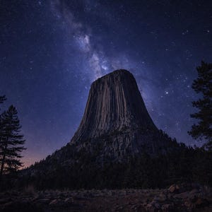 May include: A night scene of Devils Tower under a starry sky. The dark silhouette of the geological formation is prominent, with the Milky Way visible above. Pine trees frame the scene.