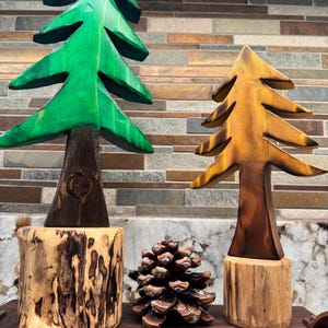 May include: Two wooden tree sculptures, one green and one brown, sit on wooden bases. A brown pine cone rests between them. The green tree has a dark brown trunk and the brown tree has a lighter brown trunk. The background is a mosaic of brown and tan tiles.