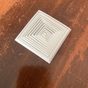 Puede incluir: Un objeto blanco de forma cuadrada con un diseño geométrico anidado. El objeto presenta múltiples capas de cuadrados progresivamente más pequeños, creando un efecto tridimensional. El artículo está sobre una superficie de madera.