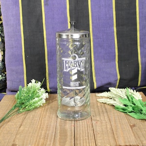 May include: A clear glass jar with a silver lid and the word "MARVY" printed on the front. The jar has a textured design and is sitting on a wooden surface. Green and white floral accents are on either side.